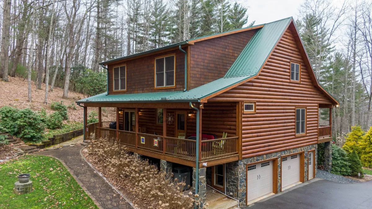 A broad view of the log cabin’s structure with its green metal roof, front porch seating, and natural landscaping. The home is surrounded by woods, emphasizing its secluded, adventure-friendly environment and easy access to nearby attractions like the river and state park.