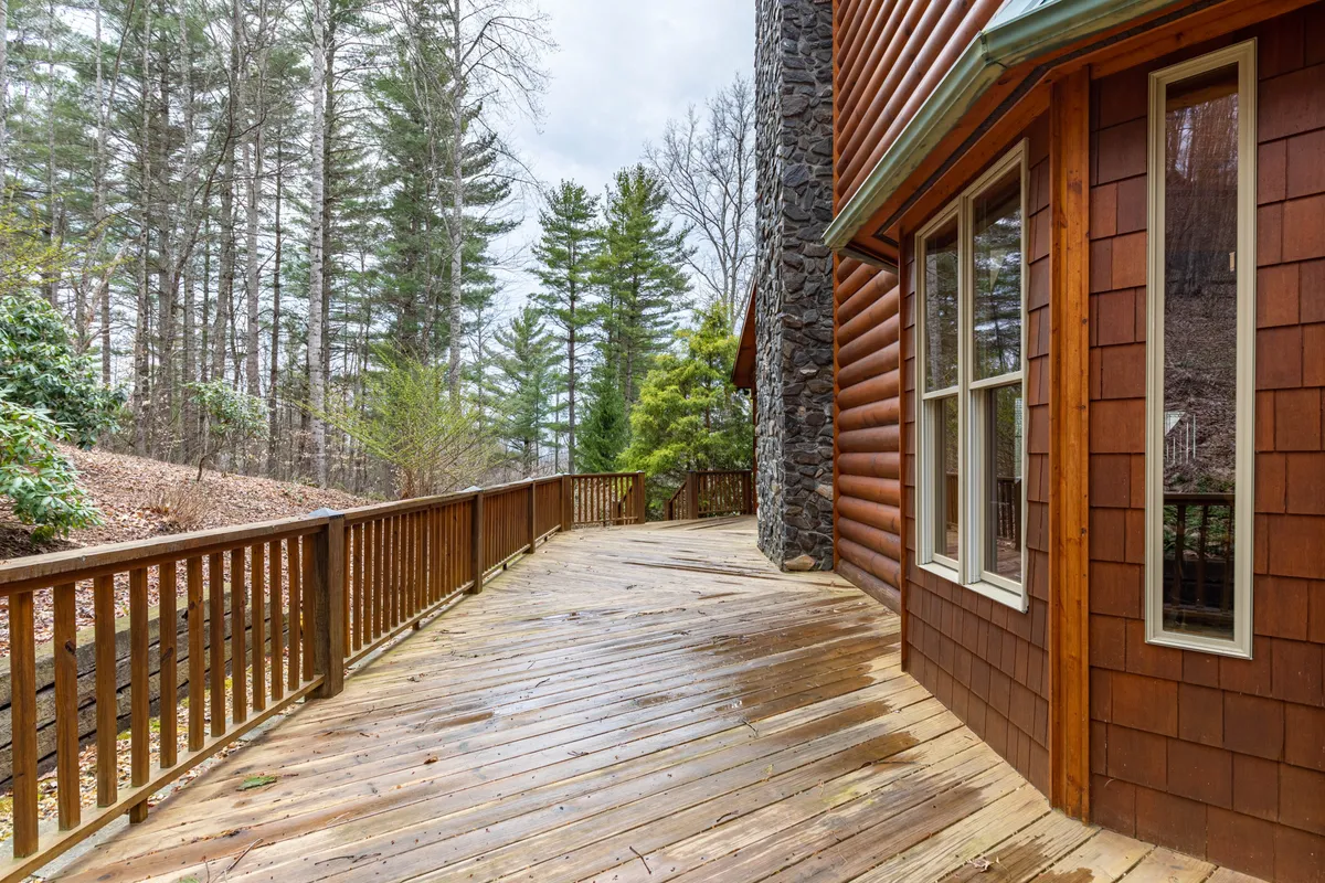 This image displays a spacious wooden deck attached to the log-sided home, with a stone wall and a tree line visible in the background. The setting is tranquil, surrounded by lush greenery, and the deck is positioned perfectly for enjoying the outdoors and nearby trails.