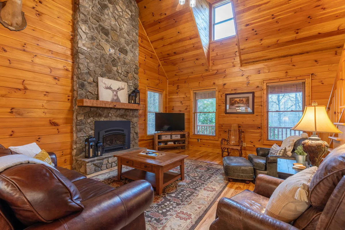 A warm, inviting living area with a stone fireplace, leather seating, a wooden coffee table, and large windows that offer views of the outdoors. The space feels open and welcoming, perfect for relaxing with family and pets after exploring outside.