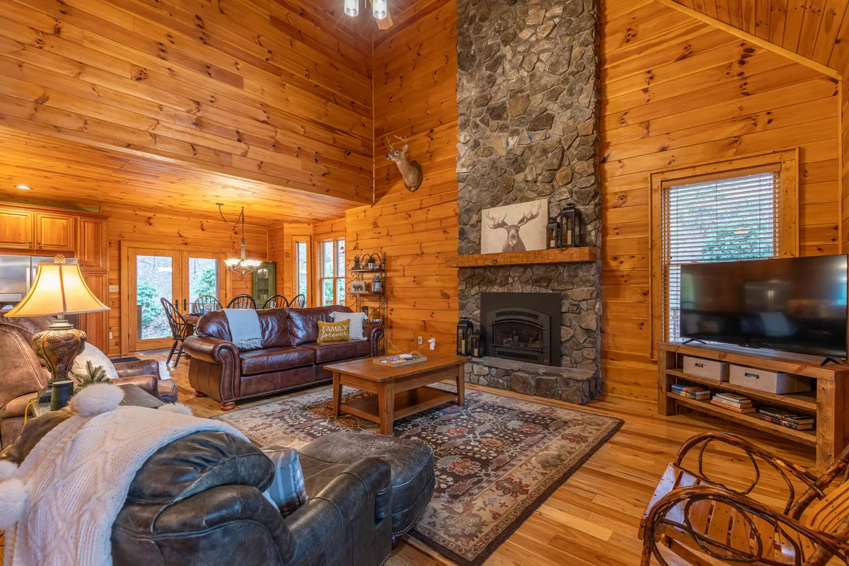 A warm living room scene anchored by a dramatic stone fireplace, wooden walls, a plush leather couch, and decorative antlers. The coffee table and large TV invite relaxation and togetherness, while rustic elements enhance the space’s charm.