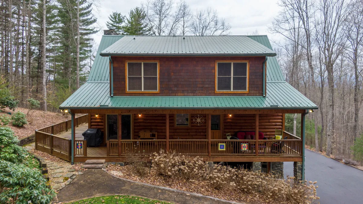 This image shows the log cabin’s inviting exterior, featuring a green metal roof, a wide front porch with comfortable seating, and a backdrop of tall, mature trees. The porch invites relaxation and hints at wildlife viewing or enjoying coffee while listening to birds.