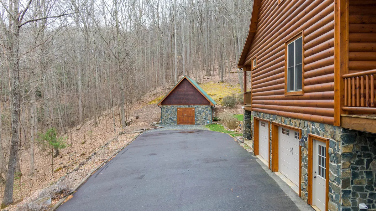 This image features the log cabin structure with a distinctive stone garage, surrounded by dense woods. The perspective highlights the cabin’s rustic charm and the sense of seclusion, inviting thoughts of wildlife observation and access to nearby trails.