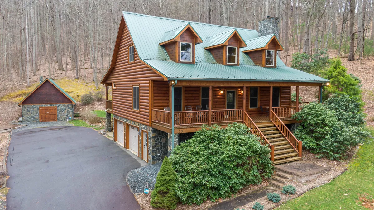 The hero image shows a charming log cabin home with stone accents, a wide welcoming front porch, and a tree-lined backdrop, all set within a peaceful driveway. The setting evokes a serene and cozy atmosphere, perfect for family gatherings and enjoying nature.