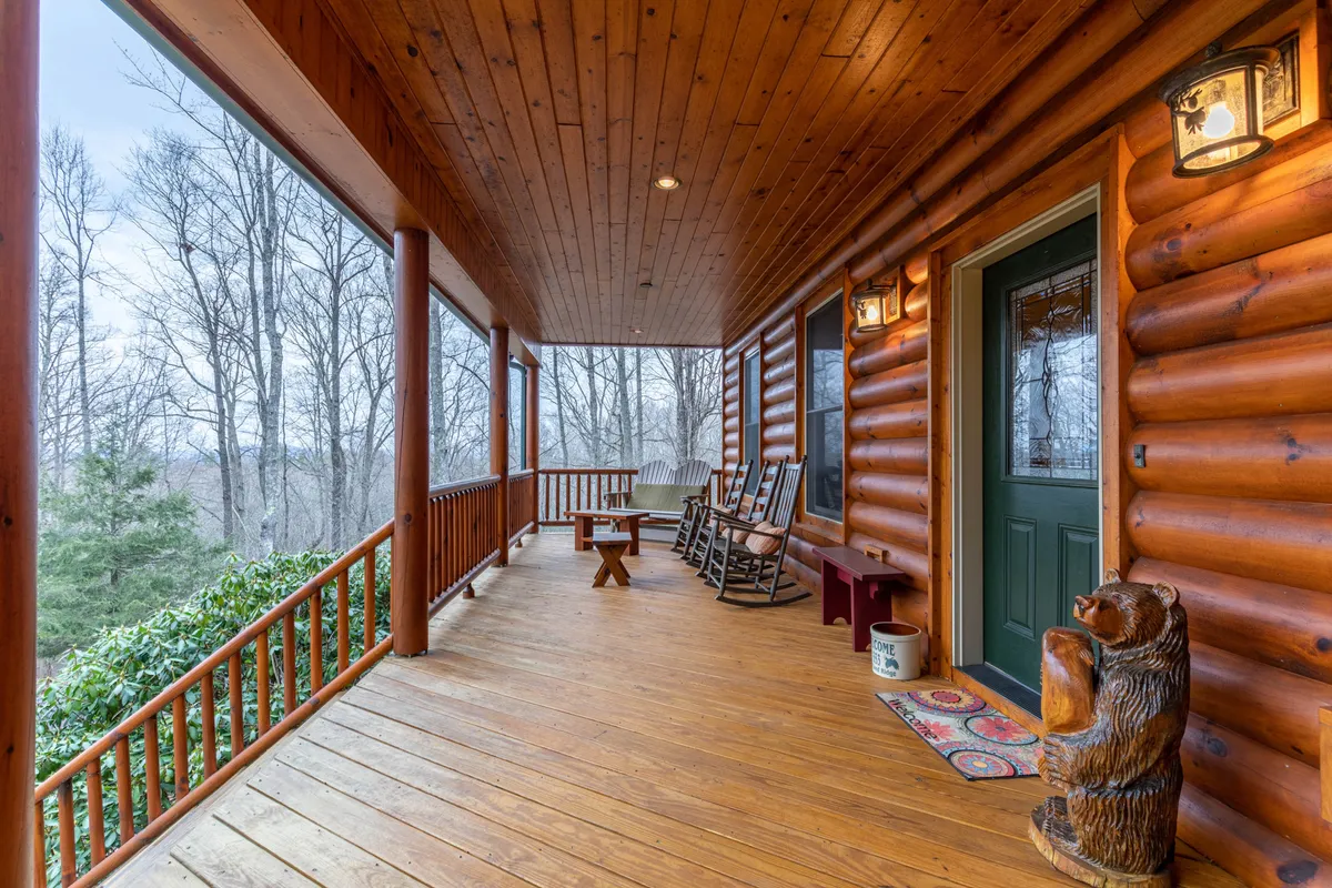 This image captures the inviting wraparound porch with classic rocking chairs, a wooden railing, and a charming decorative bear statue by the front door. The porch is bathed in soft light, hinting at sunrise or sunset, and surrounded by the sounds and sights of nature, perfect for relaxing or socializing outdoors.